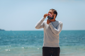 Young man in a medical mask in the color of the American flag that protects him from the virus is posing and touching the mask by his hands. Global problem concept.