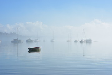 Obraz premium Lonely dinghy anchored on flat calm water with boats moored in background in morning fog.