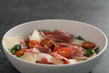 Salad with prosciutto, tomato, arugula leaves and parmesan cheese in white bowl on concrete surface
