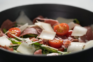 Salad with prosciutto, tomato, arugula leaves and parmesan cheese in black bowl closeup