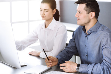 Obraz premium Friendly smiling businessman and woman working with computer in modern office. Headshot at workplace. Teamwork, partnership and business concept
