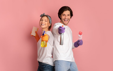 Smiling couple holding spray detergents, standing on pink background in studio