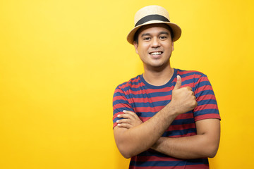 Portrait of cheerful asian man in basic clothing Casual wear red striped T-shirt and wear a hat smiling and show giving thumbs up at camera with showing success. isolated over yellow background. 