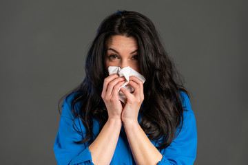 Mature woman sneezes into tissue. Isolated on grey studio background. Adult lady is sick, has a cold or allergic reaction. Coronavirus, epidemic 2020, illness concept.