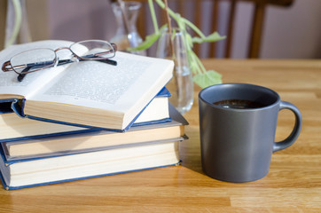 A stack of books lie on a wooden table. Nearby is a cup of coffee. horizontal background
