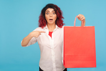 Portrait of amazed shopaholic hipster woman with fancy red hair pointing big packages and looking shocked, pleasantly surprised by successful weekend shopping. studio shot isolated on blue background