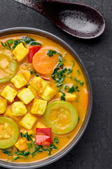 A Yellow Veg Thai Curry with Tofu and vegetables in black bowl at dark slate background. Vegetarian Thai Curry with tofu, zucchini, bell pepper, spinach, carrot. Thai Food. Indian vegetable curry