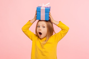 Portrait of astonished little girl holding gift box on head with expression of much surprise and...