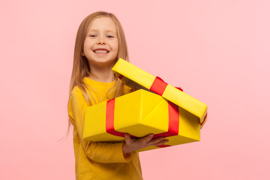 Child satisfied with good present. Portrait of charming funny little girl opening gift box and smiling to camera, celebrating birthday, Christmas holiday. studio shot isolated on pink background