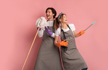 Millennial couple having fun during spring cleaning, using broom like air guitar and singing into...
