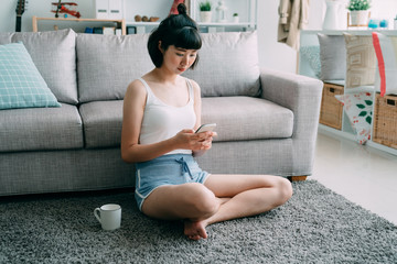 Portrait of asian chinese young woman sitting on carpet with cup of coffee. beautiful girl in summer shorts using mobile phone online relaxing on floor in modern cozy house in bright morning.