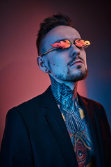 Bold and rebellious male model posing in a neon studio wearing a black tuxedo on a half-naked body, fire-shaped sunglasses and tattooed in a japanese irezumi style, looking self-assured. Vertical