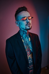 Cool and rebellious male model posing in a neon studio wearing a black tuxedo on a half-naked body, fire-shaped sunglasses and tattooed in a japanese irezumi style, looking assertive. Vertical