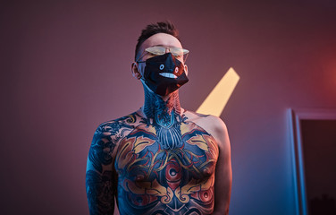 Bold and rebellious male model posing in a neon studio wearing a medical mask on his face, yellow sunglasses and tattooed in a japanese irezumi style of a half-naked body, looking self-assured.