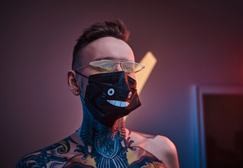 Bold and rebellious male model posing in a neon studio wearing a medical mask on his face, yellow sunglasses and tattooed in a japanese irezumi style of a half-naked body, looking self-assured.