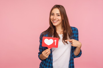 I like this content! Charming positive girl in checkered shirt smiling playfully and holding heart...