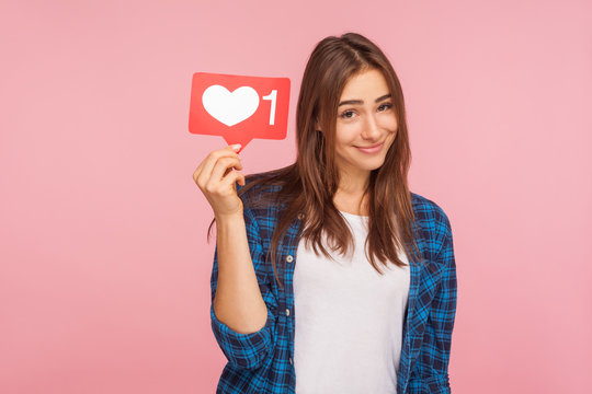 Portrait of cute positive girl in checkered shirt smiling playfully and holding social media like icon, heart button to enjoy trendy interesting content. indoor studio shot isolated on pink background