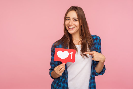 I like this content! Charming positive girl in checkered shirt smiling playfully and holding heart icon, social media button to enjoy trendy interesting blog. studio shot isolated on pink background