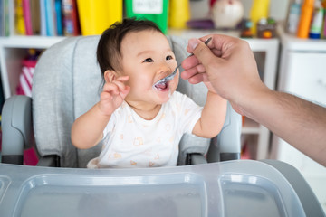 Father feed mashed food to Asian baby cute girl who sit in highchair at home. Little girl look at dad, eat yummy health food. Parent give 5 food group and high nutrition for daughter. Family concept.