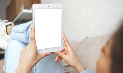 Young african woman holding in hands using digital tablet looking at mock up blank white screen for reading e book, browsing internet, watching movie or checking social media relaxing on sofa at home.