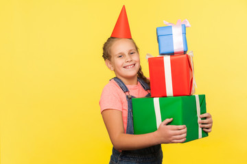 Portrait of glad little girl with funny cone holding lot of gift boxes and smiling to camera, enjoying many presents, perfect birthday party for child. indoor studio shot isolated on yellow background