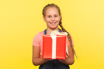 Portrait of kind merry little girl with braid holding gift box and smiling to camera, child enjoying big birthday present, Christmas holiday surprise. indoor studio shot isolated on yellow background