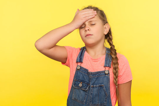 Portrait of forgetful sad little girl with braid in denim overalls gesturing facepalm with hand on forehead and expressing sorrow, blaming herself for bad memory. indoor studio shot, yellow background