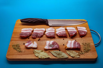 In the horizontal photo on a blue background on a Board made of wood is fresh sliced meat together with herbal seasonings for meat, Bay leaf, black pepper. Next to it is a knife with a black handle