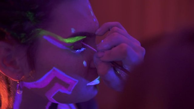 Making up a Girl with Dreadlocks in Neon UF Light. Model Girl with Fluorescent Creative Psychedelic MakeUp, Art Design of Female Disco Dancer Model in UV, Colorful Abstract Make-Up. Dancing Lady