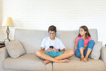 Happy boy playing with smartphone next to his bored sister looking at him