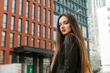 Portrait beautiful girl with long hair on background of modern urban architecture,looking in camera with a serious face.Beautiful businesswoman in black jacket posing at camera on cityscape background