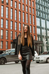 Fototapeta na wymiar Beautiful young girl in stylish formal clothes walking down the street against the backdrop of modern architecture, smiling and looking down. Vertical street photo of happy lady in office clothes