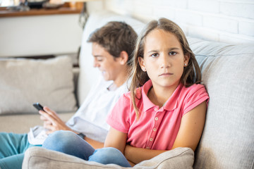 Happy boy playing with smartphone next to his sad sister looking at camera