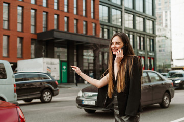 Fototapeta na wymiar Happy busy businesswoman in casual clothes talking on the phone and catches a hand gesture. Smiling formal-dressed lady communicates on the phone and stops a taxi against a cityscape.