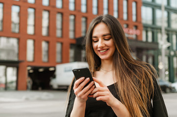 Fototapeta premium Happy lady in formal wear uses a smartphone and looks at a smartphone screen with a smile on her face. Cute smiling businesswoman communicates in a smart phone while walking through the city.