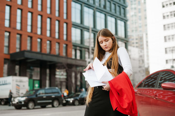 Naklejka na ściany i meble Portrait of busy student girl standing outdoors on cityscape background with laptop and notes in hands, confused looking for right piece of paper. Girl nerd is studying the way to the university.