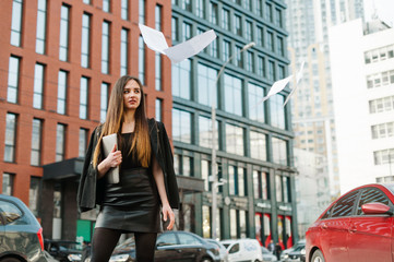 Fototapeta na wymiar Smiling office worker girl in dark formal wear tosses papers up on cityscape background, holds laptop in hands. Cheerful girl got rid of papers on the street with a smile on her face. Copy space
