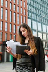 Portrait of attractive businesswoman in office clothes stands on cityscape background with laptop and papers in hands, focuses on business papers. Vertical photo. Business concept.