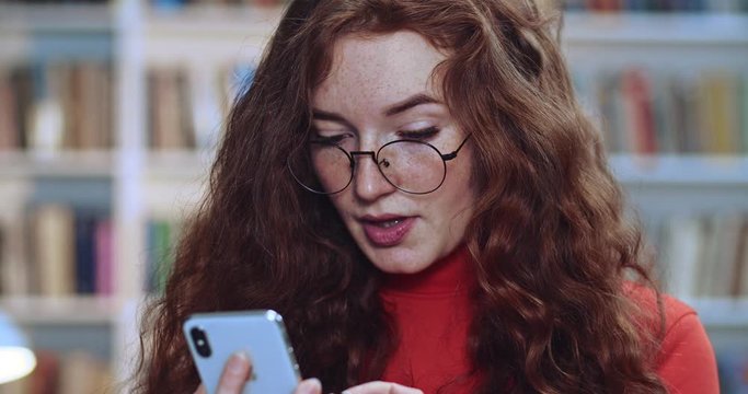 Portrait of cute red head student with long curly natural hair freckles and glasses serfing at Internet using her black phone in library. Bookcase in background.