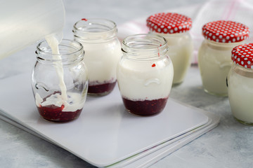Filling glass jars with a milk mixture to make yoghurt at home. Raspberry jam at bottom of jars to add sweet flavor. Organic dairy product, healthy eating and sustainable concept