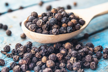 Dry black pepper in a wooden spoon near black pepper scattered on blue boards.