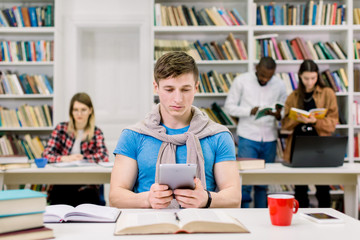 Front view of concentrated young smart guy student sitting at the table in library and working on tablet pc. Multiethnic students studying together standing on the bookshelves background