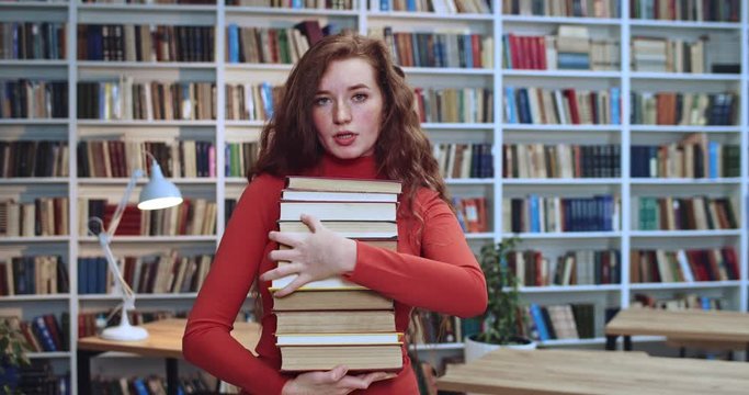 Portrait of sad red head woman with long curly natural hair and freckles joyless looking at camera and carrying books in library. Bookcase in background.