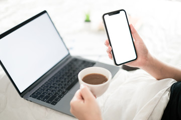 Young woman hand holding blank screen phone and coffee cup in the bed room, Work form home and holiday concept with blank screen phone and laptop computer, supplies