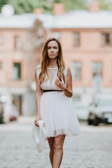 Outdoor fashion portrait of young beautiful serious lady wearing white dress, stylish straw wide brim hat, posing on street. City