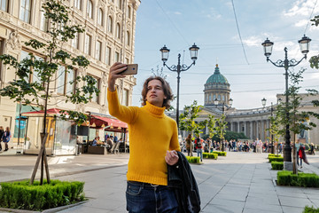 The handsome men does selfie, Look from outside, he dressed in a yellow sweater, a black raincoat or jacket is his hands, Bolshaya Konyushennaya street and Kazan Cathedral on background, sunny day