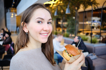 Burger in craft paper in hands and the girl looks at the camera.