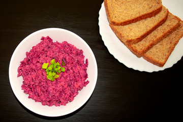 Tasty beet salad in a white bowl with brown bread on dark wooden table. Beetroot with cheese, sour cream and green onion, healthy and fresh food. 