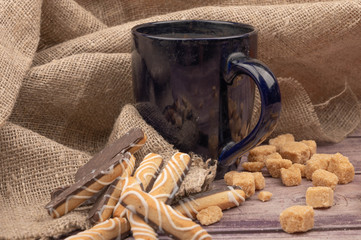 dark blue ceramic tea mug, cookie sticks with chocolate and white icing and pieces of brown cane sugar on a background of textured fabric. Close up.