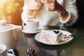 white beautiful piece of cake with blueberries on a plate in a cafe on the background of a girl with a smartphone in a blur